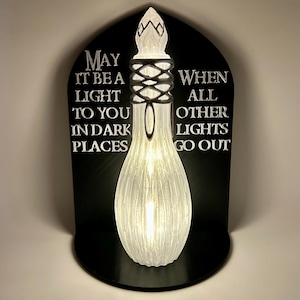 May include: A glowing, white light fixture shaped like a bottle, with a decorative top and a black base. The fixture is mounted on a black plaque with the text "MAY IT BE A LIGHT TO YOU IN DARK PLACES WHEN ALL OTHER LIGHTS GO OUT."