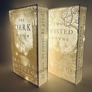May include: Two illuminated books with intricate designs. The first book, "One Dark Window," features a castle illustration. The second book, "Two Twisted Crowns," displays a tree and figure. Both books have text on the spine and cover.