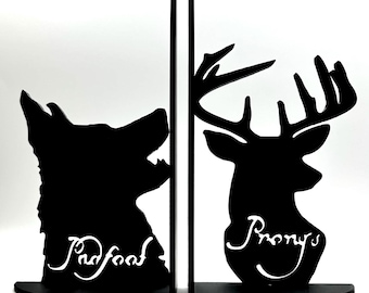 May include: Black metal bookends featuring a wolf and a deer silhouette. The wolf bookend has the word "Padfoot" in white script, and the deer bookend has the word "Prongs". These decorative bookends are designed to hold books upright.