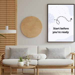 May include: A black framed print with the text "Start before you're ready" is displayed above a white sofa. The room has wooden features, including a round coffee table and a slatted wall panel. Decorative plants add a natural element to the space.