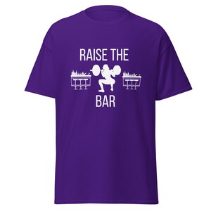 May include: Purple t-shirt with a white graphic of a person lifting a bar with two bar stools on either side of the bar. The text "Raise the Bar" is printed on the shirt.