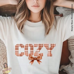 May include: Cream-colored t-shirt with the word "COZY" in a plaid pattern and a brown bow graphic. The text "Simply Shan Nicole Creations" is printed vertically on the right side of the image.