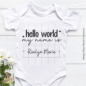 May include: White baby onesie with black text that reads "hello world my name is" with a blank space for a name and the name "Raelyn Marie" written in the space.