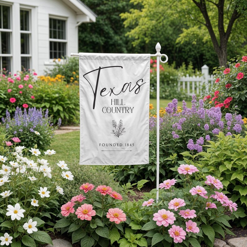 May include: A white garden flag displaying the text "Texas Hill Country" in black script, with a floral design and the founding year. The flag is on a white metal pole, set in a garden with colourful flowers.