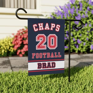 May include: A navy blue garden flag with the word "CHAPS" in red and white lettering above the number "20" in a red and white outlined font. The word "FOOTBALL" is below the number, and the name "BRAD" is at the bottom.