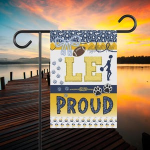 May include: A rectangular garden flag featuring a blue and white leopard print top border, a large gold letter LE, a brown football, a blue bow, a cheerleader silhouette, and the word PROUD in dark blue glittery text over a gold background.