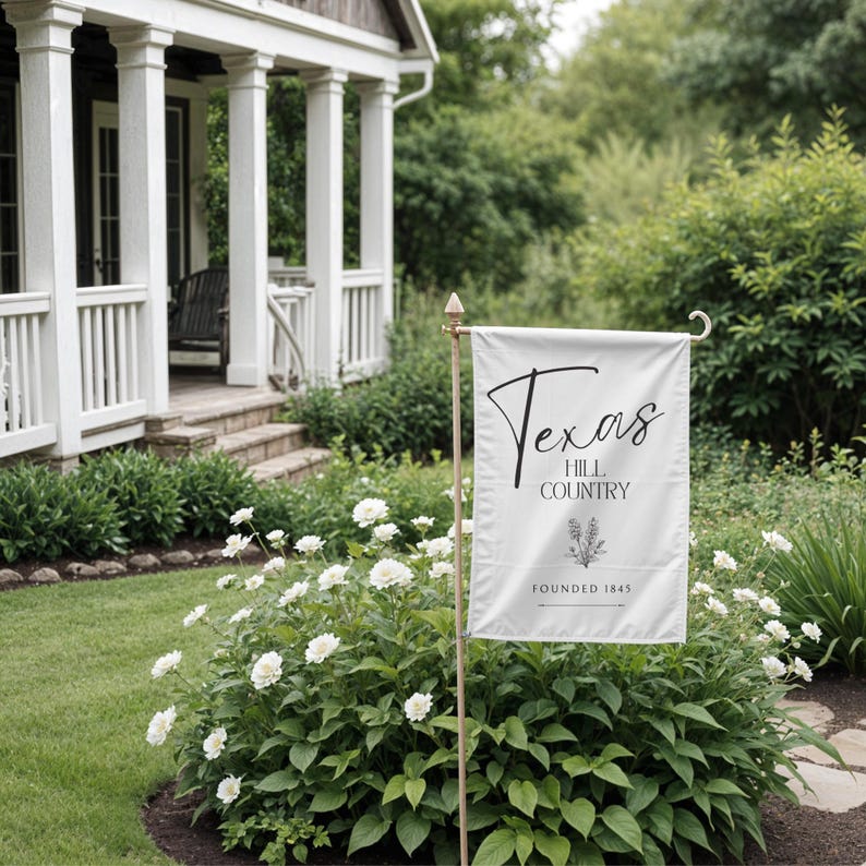 May include: A white garden flag with the words "Texas Hill Country" in black script. The flag is displayed on a gold-coloured pole in a garden with white flowers and green foliage. The words "Founded 1845" are also on the flag.