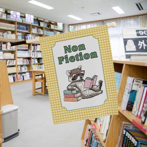 May include: A decorative flag with a raccoon wearing glasses and reading a book, with the words "Non Fiction" in green. The flag has a yellow and white checkered border and is displayed in a library setting.