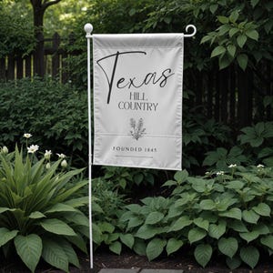 May include: A white garden flag with the words "Texas Hill Country" in black script and a floral illustration. The flag is mounted on a white metal pole and reads "Founded 1845". The flag is displayed outdoors among green foliage.