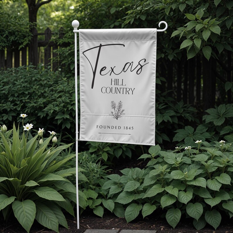 May include: A white garden flag with the words "Texas Hill Country" in black script and a floral illustration. The flag is mounted on a white metal pole and reads "Founded 1845". The flag is displayed outdoors among green foliage.