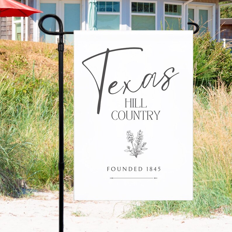 May include: A white garden flag featuring the text "Texas Hill Country" in black script. Below the text is a floral illustration and the words "Founded 1845". The flag is on a black metal stand.