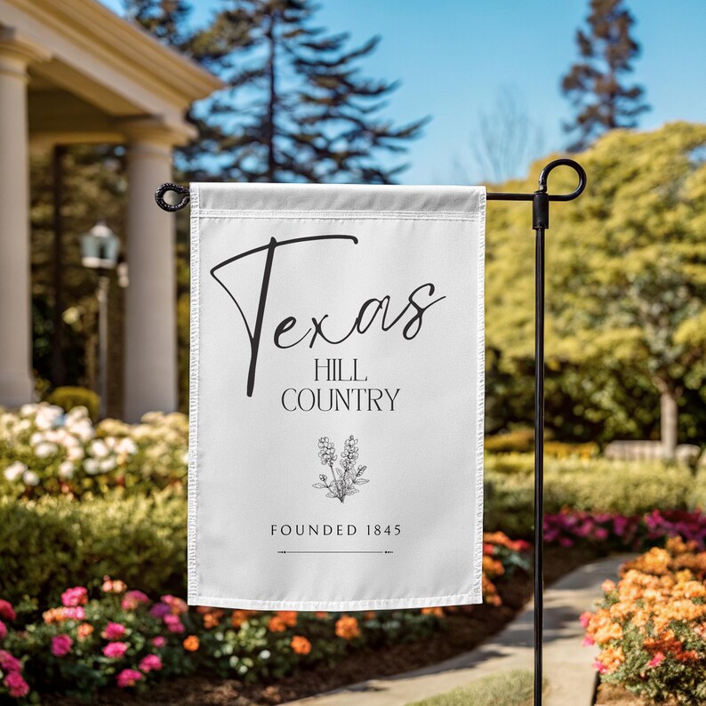 May include: A white garden flag featuring the words "Texas Hill Country" in black script and a floral illustration. The flag also includes the text "Founded 1845". The flag is displayed on a black metal stand in a garden.