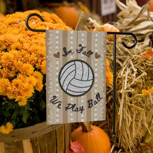May include: A decorative garden flag with a volleyball design. The flag has a tan background with vertical stripes and the words "In Fall We Play Ball." The volleyball is gray and black. The flag is displayed on a black metal stand, with orange flowers and pumpkins in the background.