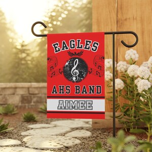 May include: A red garden flag with the text "EAGLES" and "AHS BAND" in white block letters. A musical note and treble clef are in the center. The name "AIMEE" is displayed at the bottom in a white and black border.
