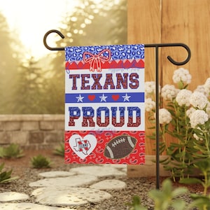 May include: A garden flag with a patriotic design. The flag features the words "TEXANS PROUD" in red, white, and blue, along with stars, hearts, and a football graphic. The flag is displayed on a black metal stand.