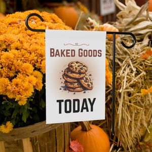 May include: A white garden flag with the words "Baked Goods Today" and an illustration of chocolate chip cookies. The flag is displayed on a black metal stand, surrounded by orange flowers, pumpkins, and hay.