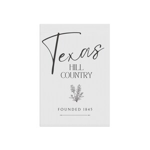 May include: A white rectangular sign featuring the text "Texas Hill Country" in a black script font. Below the text is a floral illustration and the words "Founded 1845". The sign has a minimalist design.