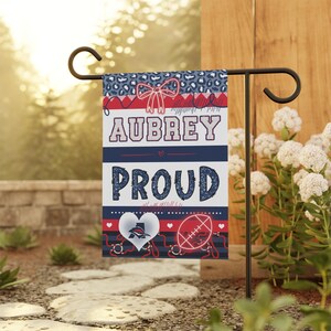 May include: A garden flag with the name "AUBREY" and the word "PROUD" in a decorative font. The flag features a bow, hearts, and a football design, with a color scheme of red, white, and blue. The flag is displayed on a black metal stand.
