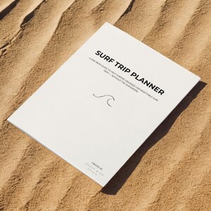 Könnte beinhalten: Ein weißes Surf Trip Planner-Buch mit schwarzem Text und einer Wellengrafik. Auf dem Cover steht "SURF TRIP PLANNER" und "A ONE-WEEK GUIDE TO HELP SURFERS ORGANIZE UNFORGETTABLE SURF TRIPS - WITHOUT THE GUESSWORK." Das Buch liegt auf sandigem Untergrund.