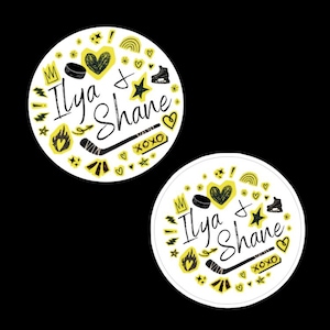 May include: Two round white stickers with black and yellow illustrations. Each sticker has the names "Ilya & Shane" in script, surrounded by hearts, stars, a hockey stick, a crown, and a hockey skate.
