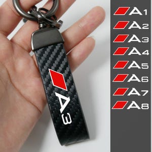 Puede incluir: Llavero negro y de fibra de carbono con un logotipo de diamante rojo y el texto blanco "A3". El llavero tiene un anillo de metal y una correa de cuero negro. Una lista de modelos de coches A1-A8 está a la derecha.