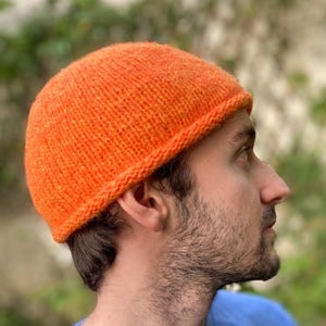 Peut inclure: Un bonnet en tricot orange vif. Le bonnet a un bord côtelé et est fait de fil texturé. La personne sur l'image porte un t-shirt bleu. Le bonnet semble être fait à la main.