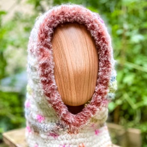 Peut inclure: Un capuchon crocheté à la main en fil crème, rose et vert. Le capuchon présente une bordure duveteuse brun-rougeâtre autour de l'ouverture du visage. L'article est présenté sur une forme de tête en bois, mettant en valeur la texture et les variations de couleurs du fil.