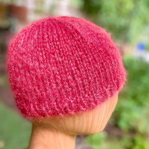 Può includere: Un berretto lavorato a mano di colore rosso lampone. Il cappello è realizzato con un filato strutturato e viene mostrato su una testa di legno. Il motivo a maglia fitta del berretto suggerisce calore e comfort.