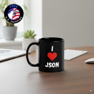 May include: Black ceramic mug with a white "I" above a red heart, followed by the word "JSON" in white. The mug has a curved handle and sits on a wooden surface. A "Free U.S. Shipping" logo is visible in the background.