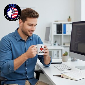 May include: A white ceramic mug with the text "I ❤️ JSON" in black and red. The mug is held by a person wearing a blue button-down shirt. The background includes a computer, desk, and office supplies.