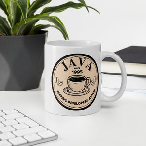 May include: White ceramic coffee mug with a black handle. The mug features a circular design with the word "JAVA" above a coffee cup illustration, the text "SINCE 1995", and the phrase "KEEPING DEVELOPERS AWAKE".