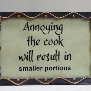 May include: A black framed sign with gold accents and a white background. The sign reads "Annoying the cook will result in smaller portions".