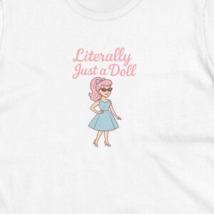May include: White t-shirt with the text "Literally Just a Doll" above a cartoon illustration of a doll wearing a blue dress and sunglasses. The doll has pink hair and is standing with her hands on her hips.