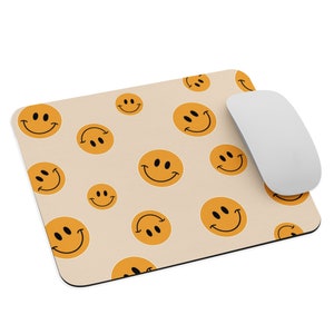 Puede incluir: Una alfombrilla de ratón beige con un patrón repetitivo de caras sonrientes naranjas. Un ratón de ordenador blanco está en la alfombrilla.
