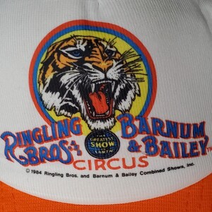 May include: A white baseball cap featuring a graphic of a tiger head inside a circular frame. Below the tiger, the text reads Ringling Bros. and Barnum & Bailey Circus, The Greatest Show on Earth, and 1984 Ringling Bros. and Barnum & Bailey Combined Shows, Inc.