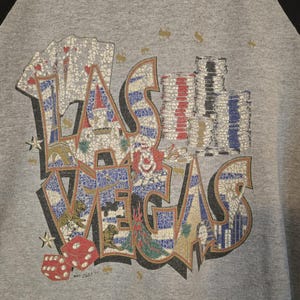 May include: Gray raglan-style shirt with a large, colorful graphic that reads "LAS VEGAS." The graphic includes playing cards, dice, poker chips, and images of tigers and landmarks. The sleeves are black.