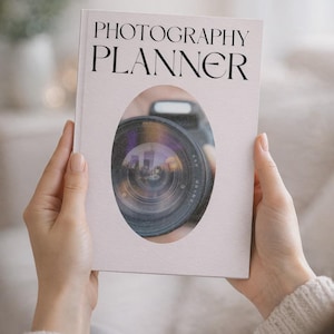 May include: A white photography planner book with black serif text reading PHOTOGRAPHY PLANNER. The cover features a central oval cutout revealing a detailed view of a black camera lens with a visible glass element and a small flash unit.