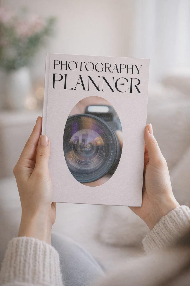 May include: A white photography planner book with black serif text reading PHOTOGRAPHY PLANNER. The cover features a central oval cutout revealing a detailed view of a black camera lens with a visible glass element and a small flash unit.