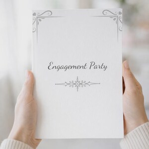 K&ouml;nnte beinhalten: Ein wei&szlig;es rechteckiges Buch mit dem in Schwarz gedruckten Text Engagement Party in der Mitte. Der Einband zeigt graue dekorative Schn&ouml;rkel an den Ecken und ein zentriertes graues ornamentales Verzierungsdesign unter dem Text.