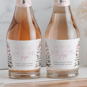 May include: Two mini bottles of sparkling wine with pink and white labels that say "Pop it when she pops". The labels also include a watercolor illustration of a snowy forest with pink bows and the text "Enjoy a toast when baby Miller arrives".