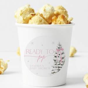 May include: White popcorn cups with a sticker that says "Ready to Pop" with a pink ribbon and a white tree design. The text "Sandra's Baby Shower" is also on the sticker.