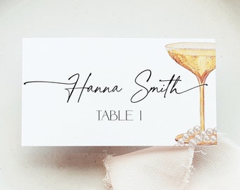 Puede incluir: Tarjeta de lugar blanca con caligrafía negra que dice "Hanna Smith" y "Mesa 1". A la derecha de la tarjeta se ilustra una copa de champán dorada con un collar de perlas.