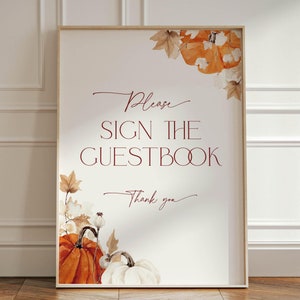 May include: A white sign with a brown wooden frame featuring a watercolor illustration of pumpkins and fall leaves. The text on the sign reads "Please sign the guestbook. Thank you."