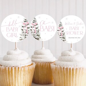May include: Three cupcake toppers with a winter theme. The toppers feature a watercolor illustration of a pine tree with pink bows and snowflakes. The text on the toppers reads "hello baby girl", "oh baby", and "Welcome to Sandra's Baby Shower December 4th, 20xx".