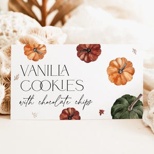 May include: A white rectangular card with the text "VANILLA COOKIES with chocolate chips" and watercolor pumpkins in orange, red, and green. The card is decorated with small leaf illustrations.