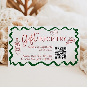 May include: A white card with a green border and a wavy edge. The text "Gift Registry" is written in red cursive. The text "Sandra is registered at Amazon." is written in black. A QR code is in the center of the card. The text "Please scan the QR code to view the gift registry." is written in black.