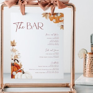 May include: A printable sign for a bar menu with a fall theme. The sign features a watercolor illustration of pumpkins and leaves in shades of orange, brown, and white. The text "The Bar" is written in a cursive font in burgundy. The menu lists beer, wine, soda, juices, and cocktails.
