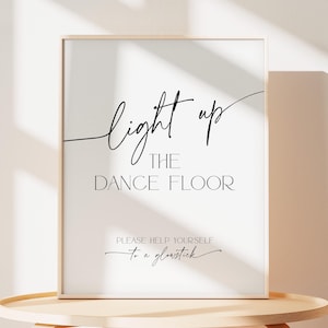 Puede incluir: Un cartel blanco con texto negro que dice "Light up the dance floor. Please help yourself to a glowstick." El cartel está enmarcado en un marco de madera clara y está sentado en una mesa de madera clara.