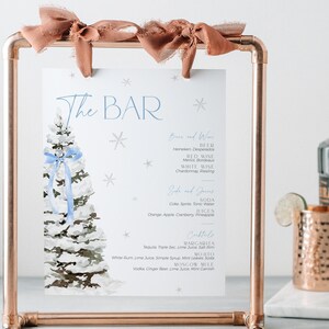 May include: A printable sign for a bar with a wintery theme. The sign features a watercolor illustration of a snowy pine tree with a blue ribbon. The sign lists the drinks available at the bar, including beer, wine, soda, juices, and cocktails. The sign is displayed on a copper pipe stand.