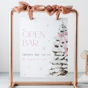 May include: A white sign with the text "Open Bar" and "Drinks are on us, the hangover is on you". The sign is decorated with a watercolor illustration of a snowy pine tree with a pink ribbon. The sign is displayed on a copper pipe stand.
